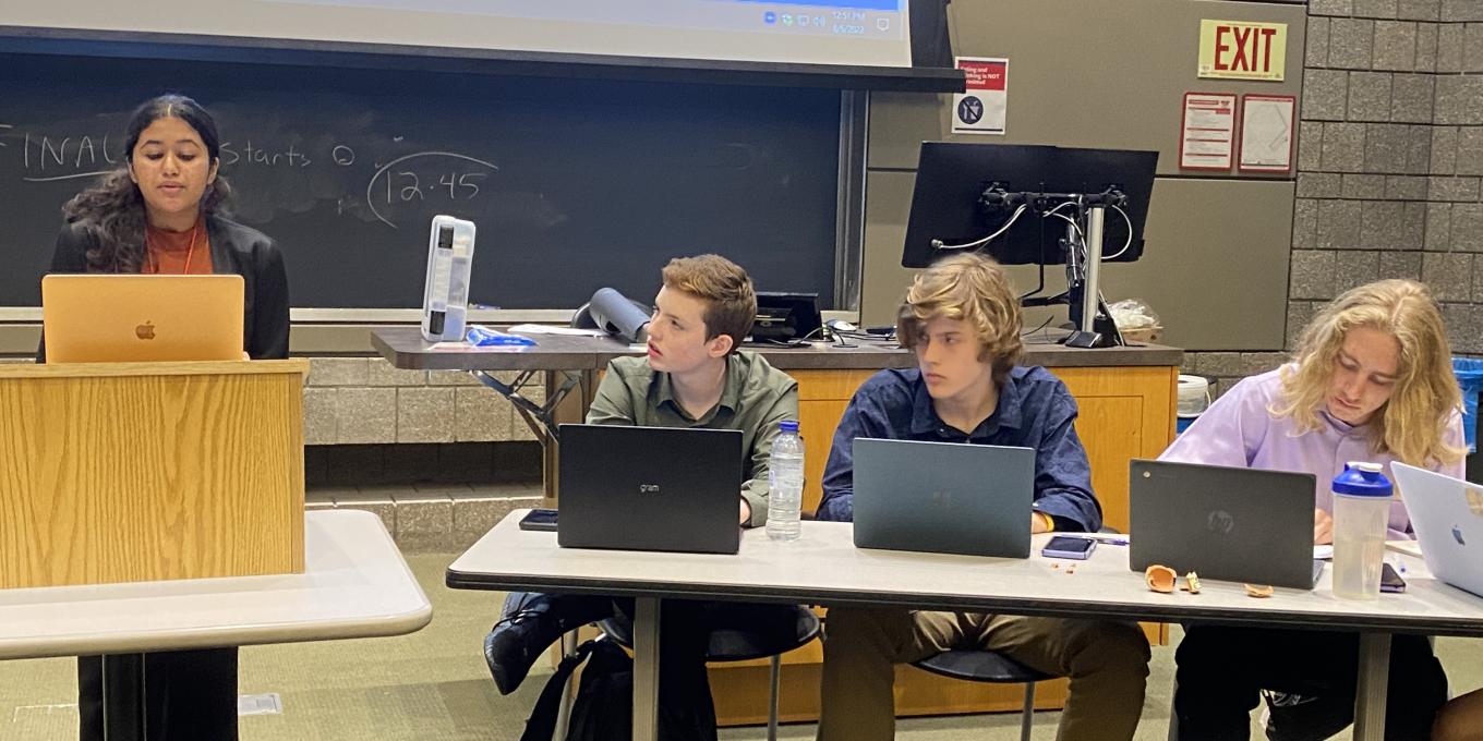 one student speaks at a podium while three students sit at laptops listening