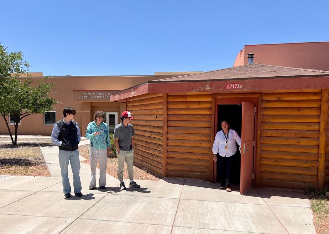 Three Cornell undergraduates stand in bright sun on a sidewalk, looking at a person standing in a hogan’s open doorway. The hogan has walls made of horizontally stacked logs and a shallow, sloped roof with a vent in the center.