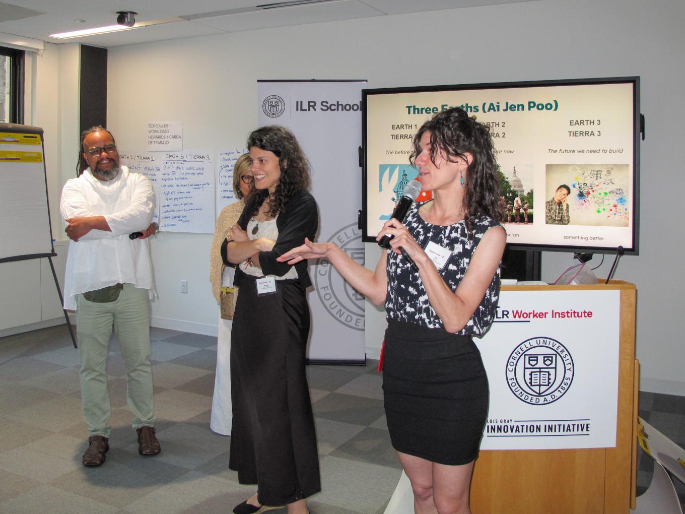 Woman standing speaking into a microphone addressing group outside of shot. Man and woman standing smiling beside her