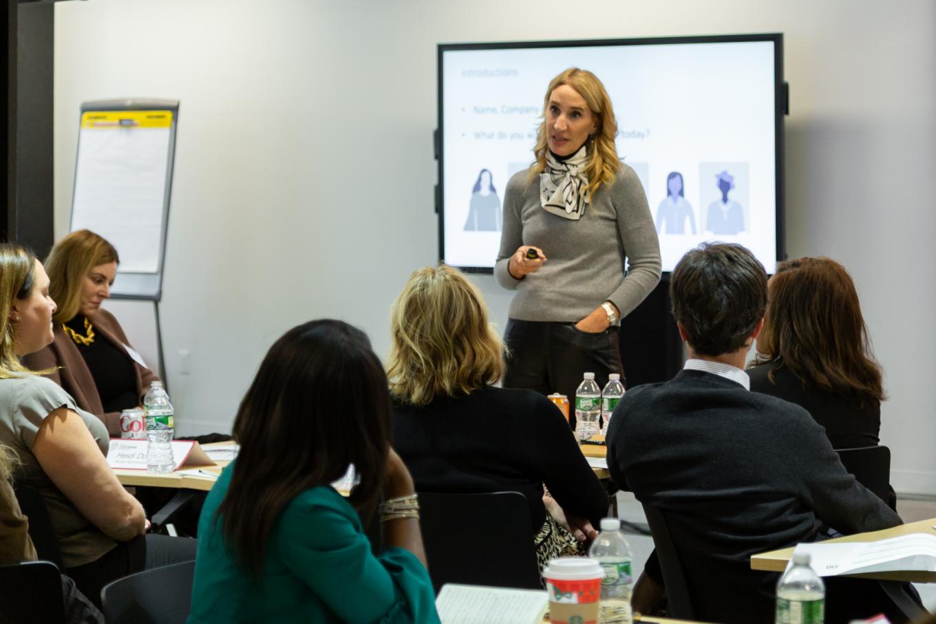 A presenter speaks to a seated group during a professional workshop, with an introductions slide displayed on a screen behind her.