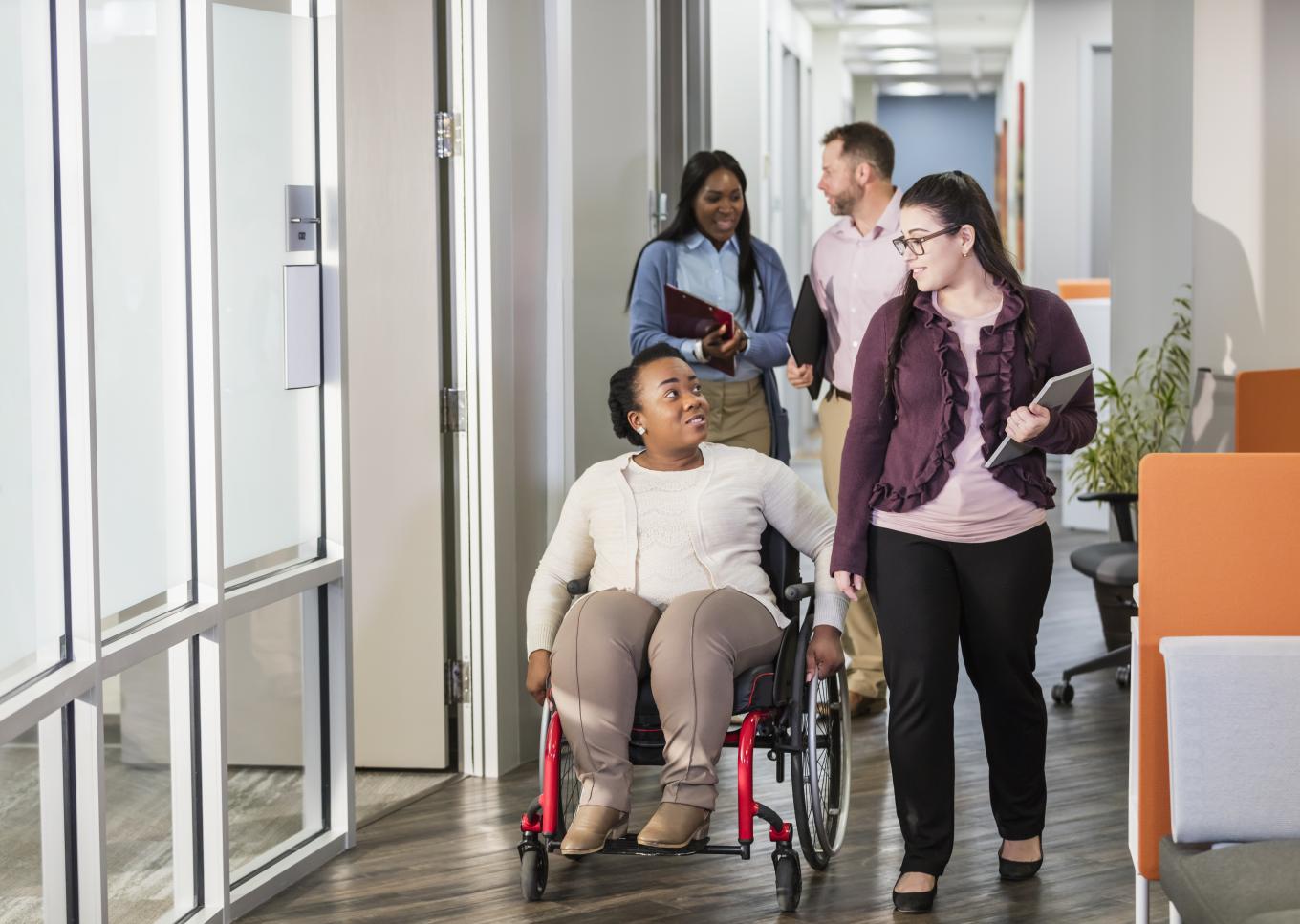 Colleagues chatting in the workplace; one is ambulating in a red wheelchair, others are walking.