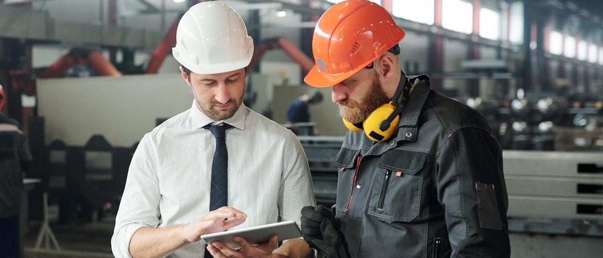 Two workers wearing hard hats. One holds a tablet and is supervising the other.