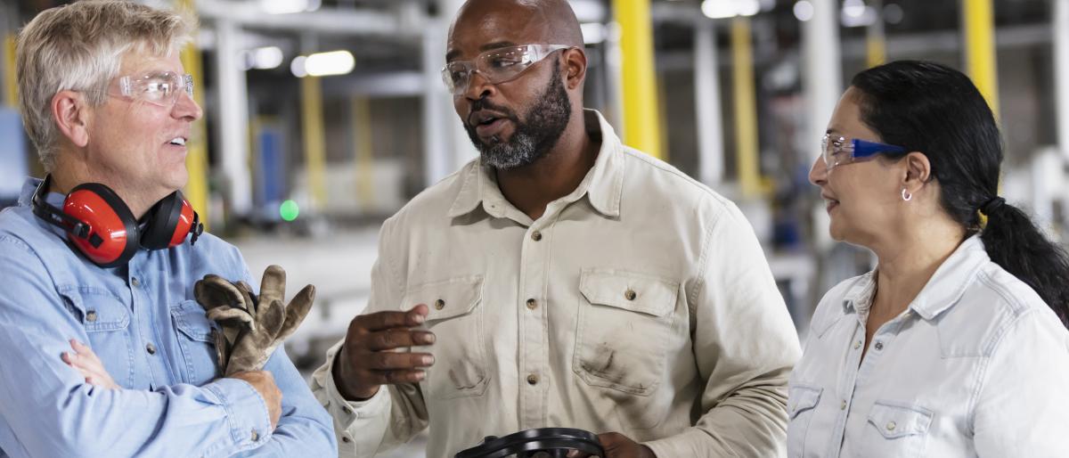 Two men and a woman having a conversation on a manufacturing floor, wearing safety glasses, ear protection and holding work gloves. 