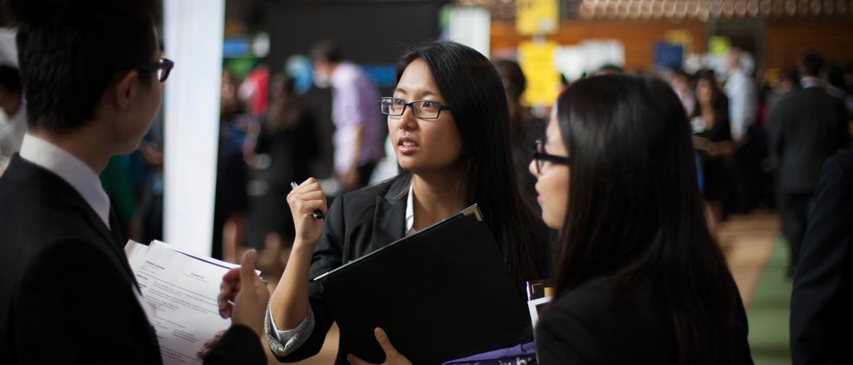 Students in professional attire speak with a recruiter while holding résumés and portfolios at a crowded career fair in Barton Hall.