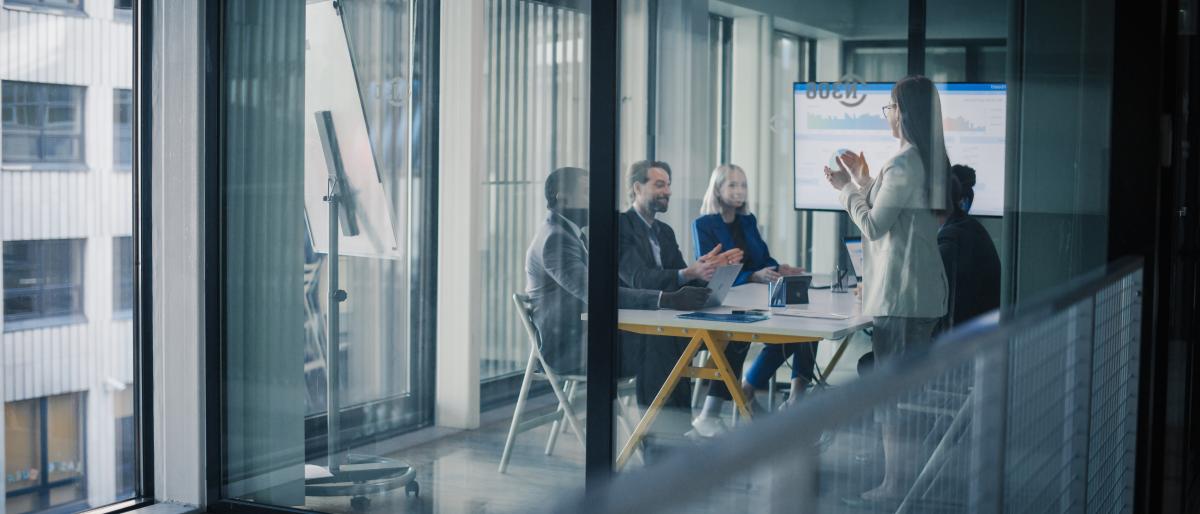 Professional presenting to colleagues during a strategy meeting in a modern glass-walled conference room.