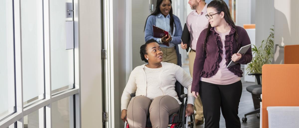 Colleagues chatting in the workplace; one is ambulating in a red wheelchair, others are walking.