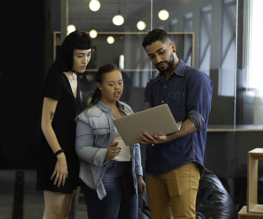 Three employees in the workplace stand together while viewing a laptop screen