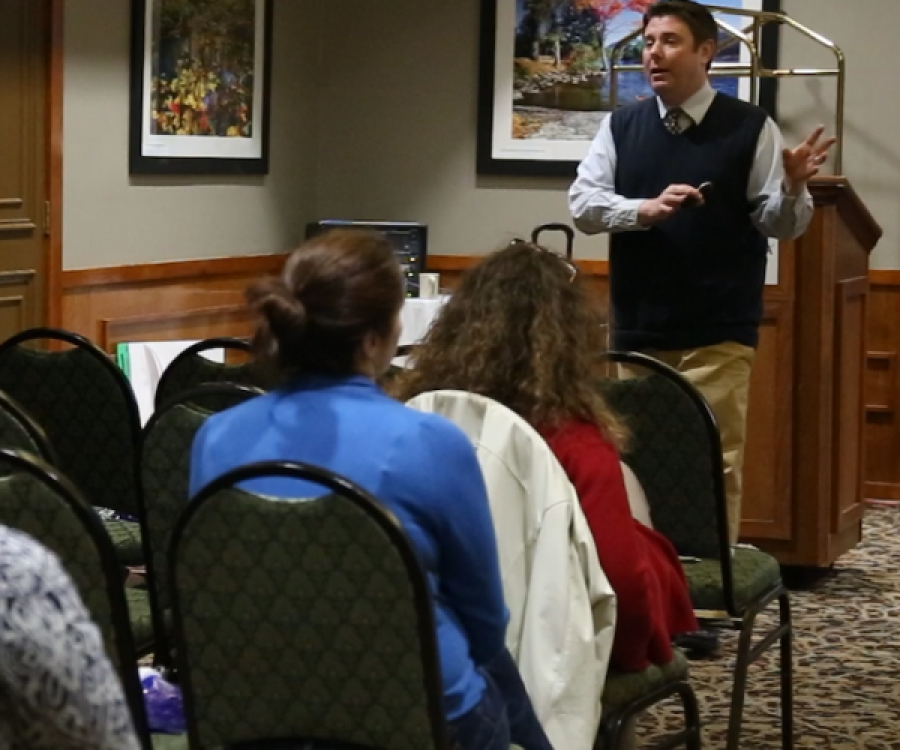 Instructor Jeffrey Tamburo wears a long sleeve light blue button up shirt with a necktie and blue sweater vest and stands in front of a podium while lecturing seated participants