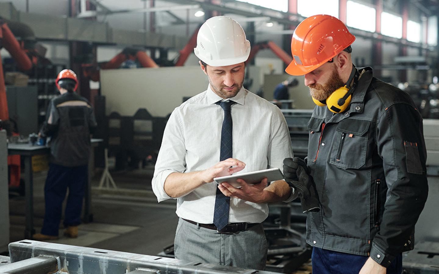 Two workers are wearing hard hats. One holds a tablet and is supervising the other.