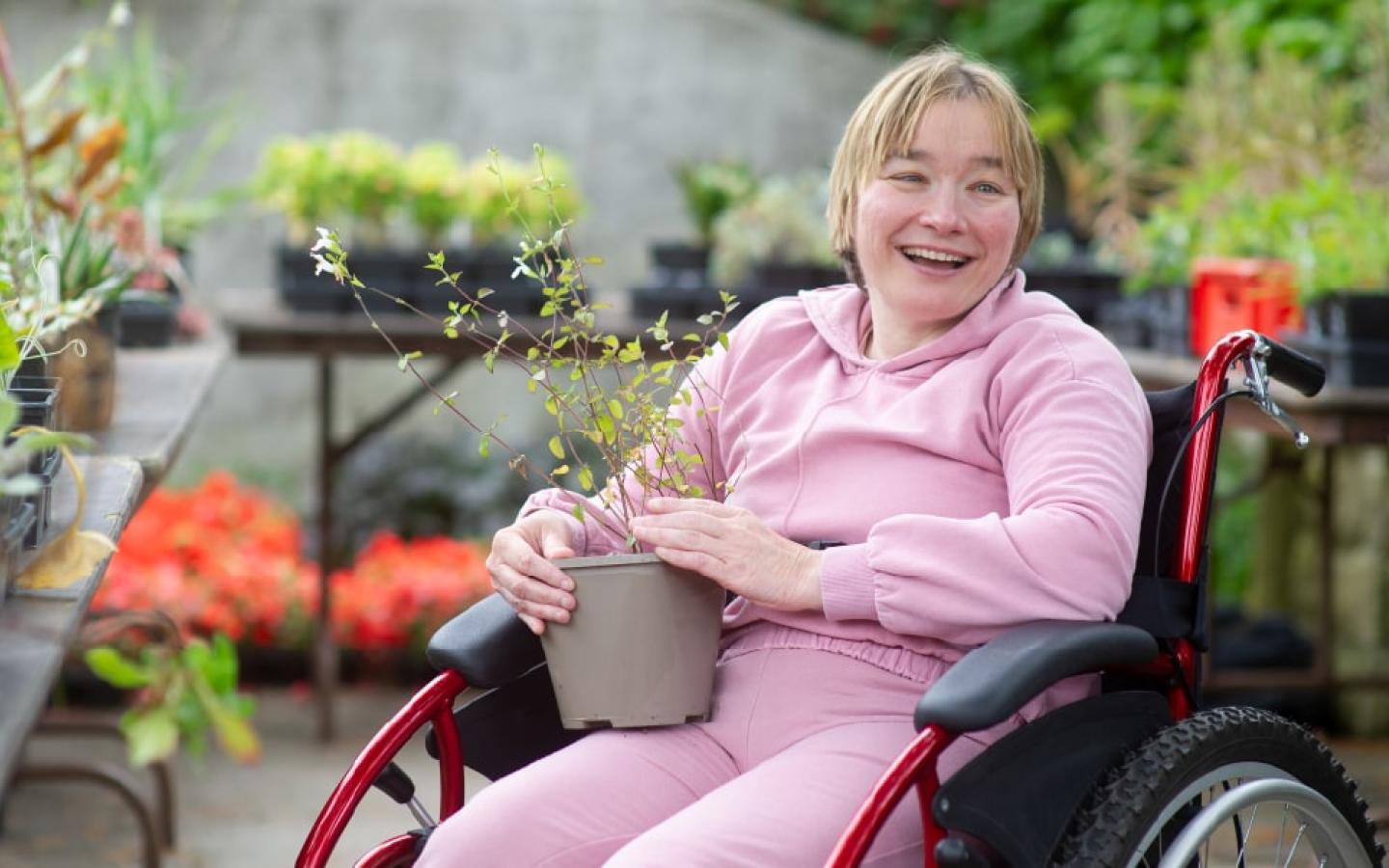 Woman in a wheelchair surrounded by greenery outdoors, holding a potted plant