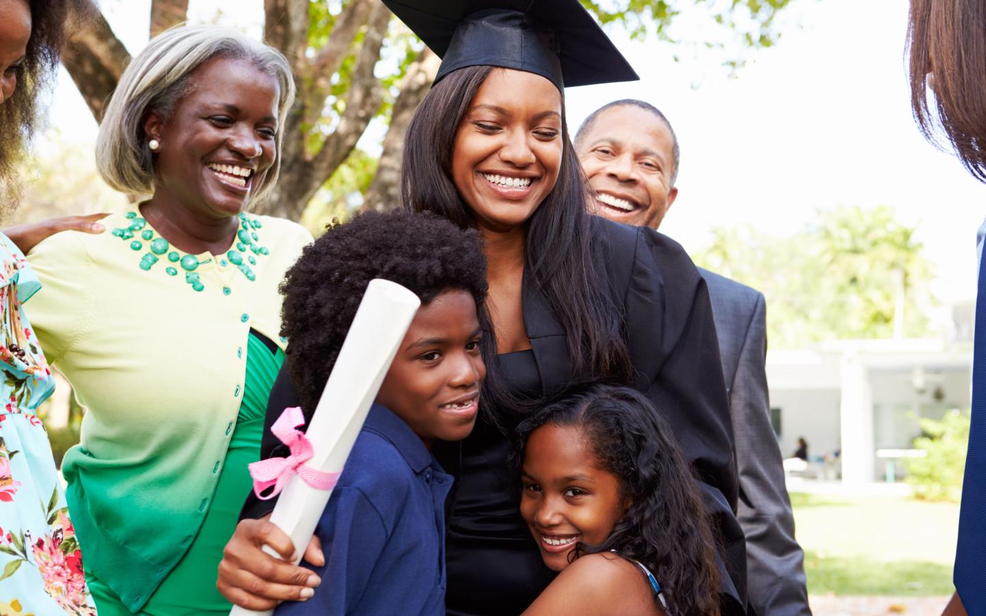 young woman in graduation attire, holds a diploma while hugging family members