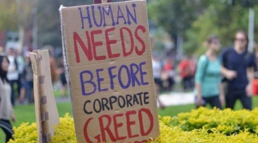 A protest sign that says, "Human needs before corporate greed." The sign is posted in a bush and people are walking in the background.