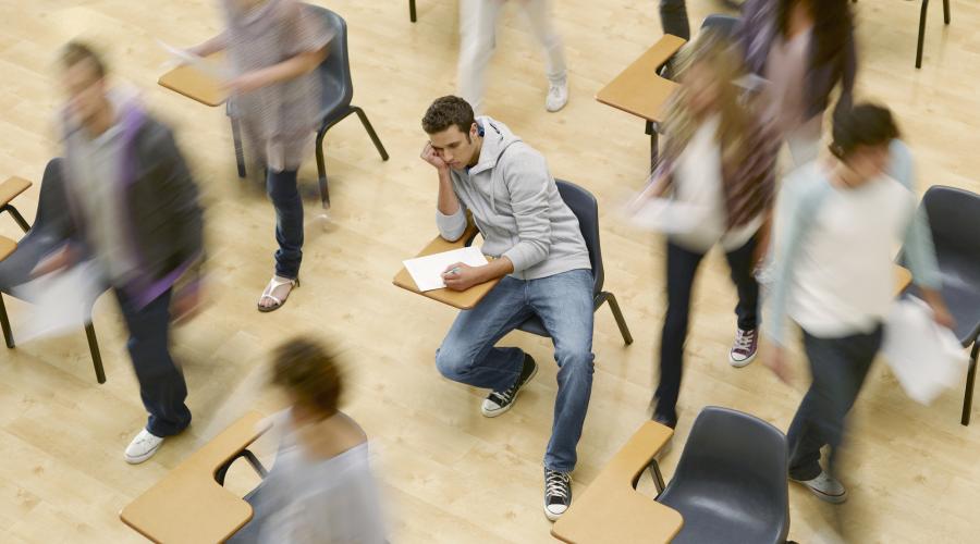 A college student sits at a desk while his classmates rush around