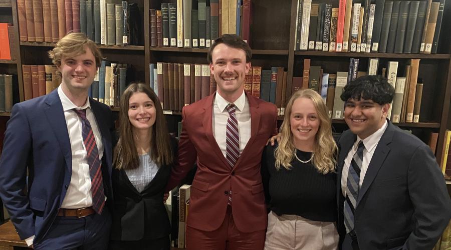 5 Cornell representatives in dress clothes stand in front of a book case