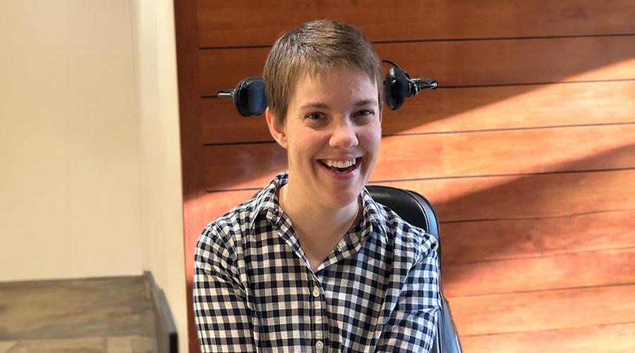 Jen Brooks, seated in her powerchair in the lobby of the Yang-Tan Institute, is smiling at the camera.
