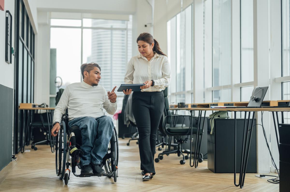 Businessman in a wheelchair is using a digital tablet while having a discussion with a colleague in the corridor of a co-working space.