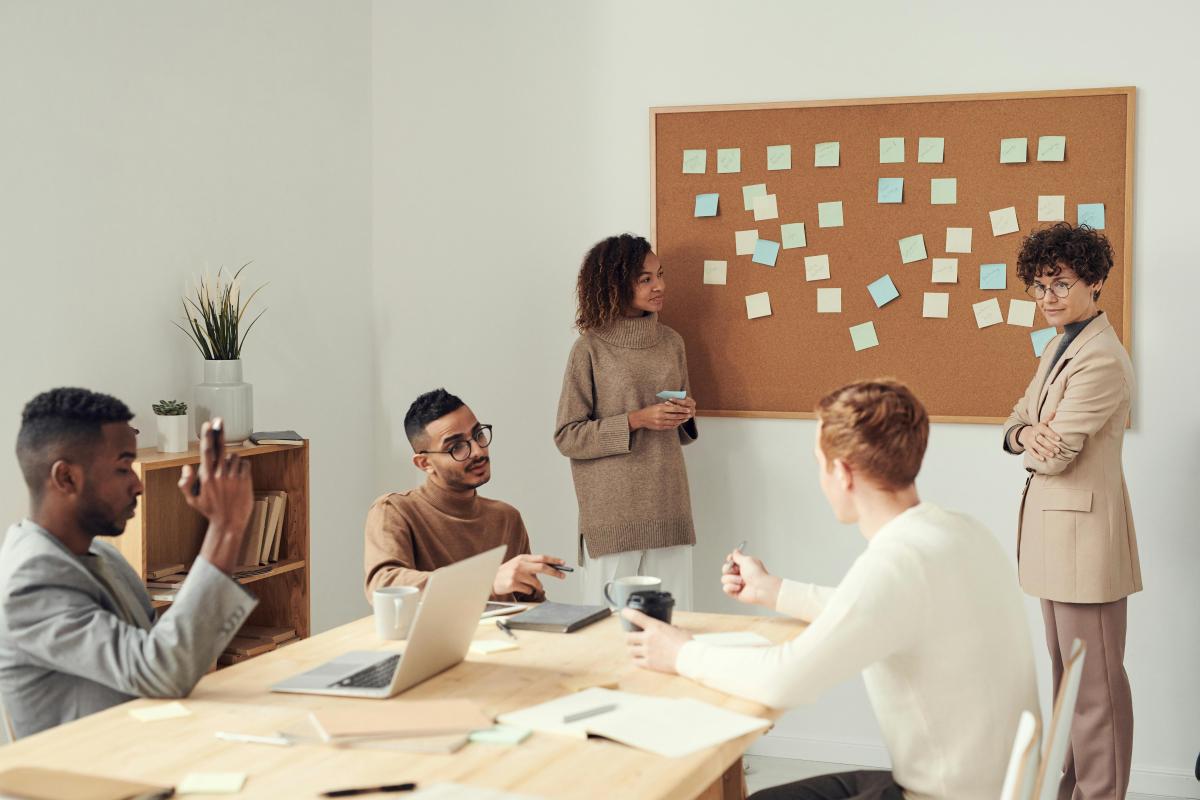 3 people sitting at a conference table looking up at 2 women presenting on cork board