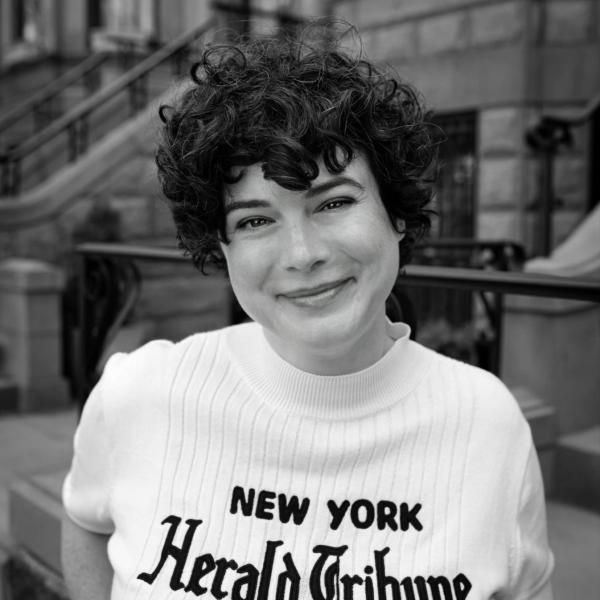 Sarah Krasley, a woman with short curly dark hair, smiles as she wears a shirt that says New York Herald Tribune in a black and white photo