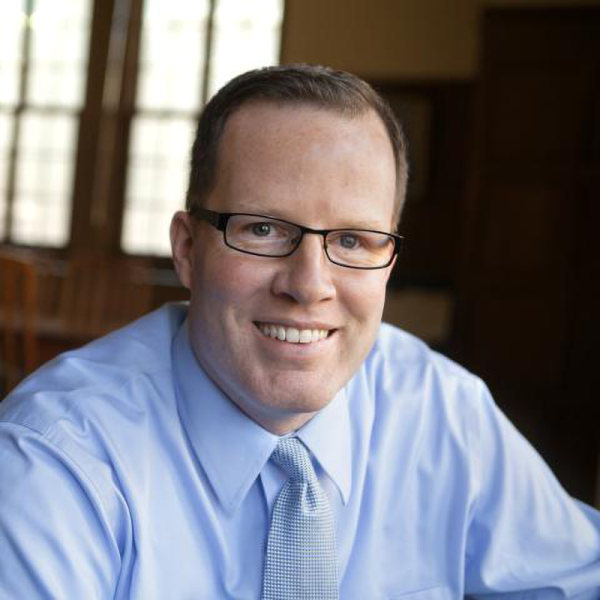 Smiling man wearing glasses, a light blue dress shirt, and a light blue tie, seated indoors near wood-framed windows.