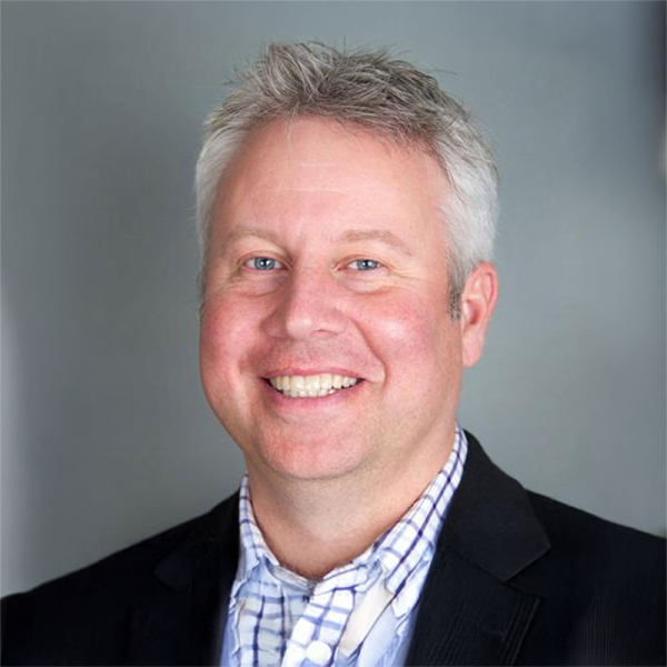 Smiling man with short gray hair wearing a black jacket over a light checkered shirt, posed against a neutral gray studio background.