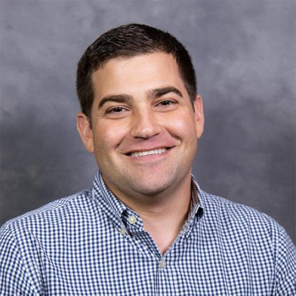Smiling man with short dark hair wearing a blue-and-white checkered button-down shirt, posed against a neutral gray studio background.