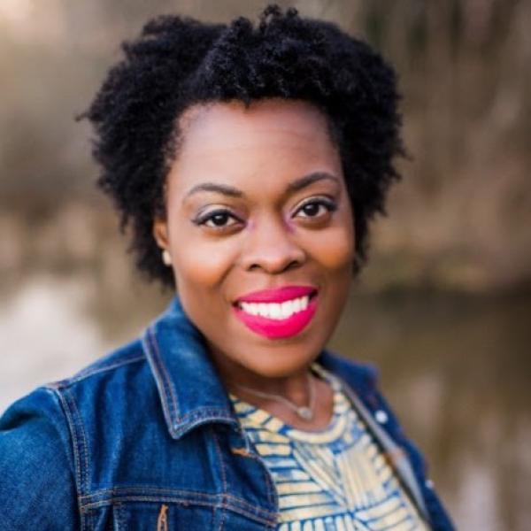 Portrait photo of a smiling person outdoors, with short natural curly hair, a denim jacket, and bright red lipstick against a softly blurred background.