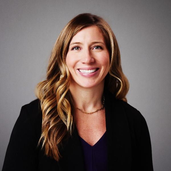Professional headshot of a smiling person with long wavy hair, wearing a dark blazer and top, photographed against a plain gray background.