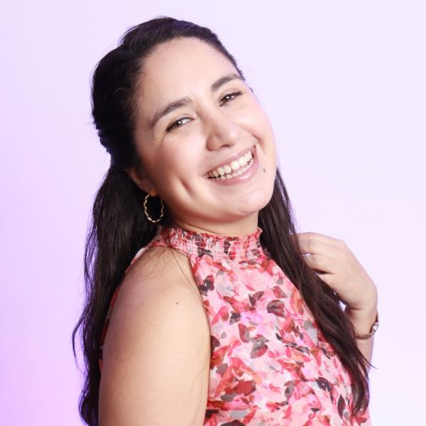Studio headshot of a smiling person with long dark hair, wearing hoop earrings and a pink floral sleeveless top, posed against a light lavender background.