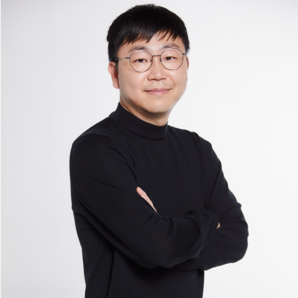 Studio headshot of a person wearing round glasses and a black long-sleeve mock-neck top, standing with arms crossed against a light gray background.
