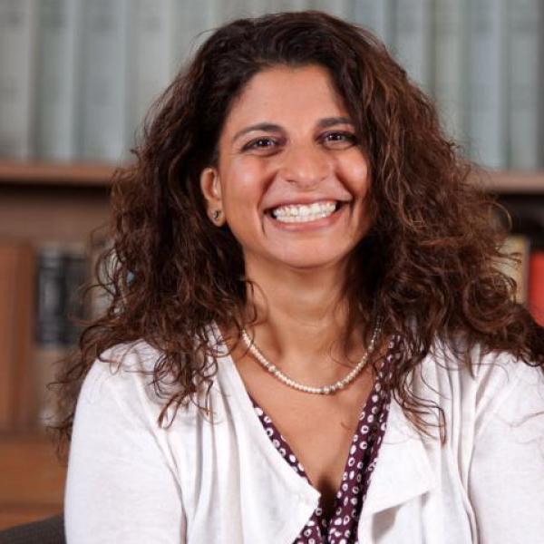 A smiling woman with curly hair sits in front of bookshelves, wearing a white cardigan and pearl necklace.
