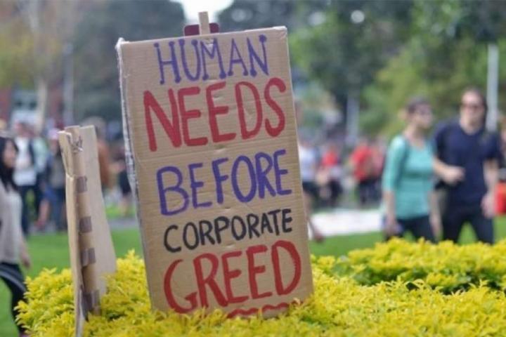 A protest sign that says, "Human needs before corporate greed." The sign is posted in a bush and people are walking in the background.