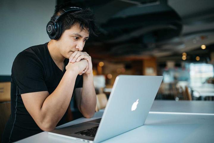 A young man in a T-shirt sits at a laptop wearing headphones.