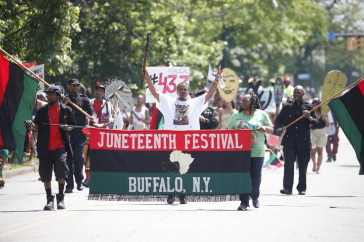 A group marches behind a sign that reads "Juneteenth Festival Buffalo, NY with an image of the African continent. Marchers hold up Black Liberation Flags and large cutout drawings of African masks. 