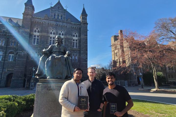 Two Cornell debaters stand with coach in front of a statue at Georgetown 