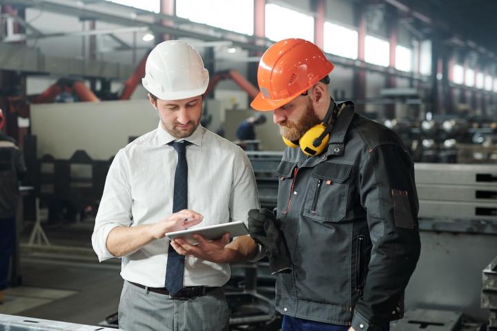 Two workers wearing hard hats. One holds a tablet and is supervising the other.
