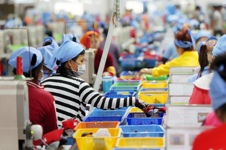 Cambodian workers are seen in a local footwear manufacturing plant, with containers of materials in multiple colors sitting in front of workers wearing bandanas and many wearing masks as well.