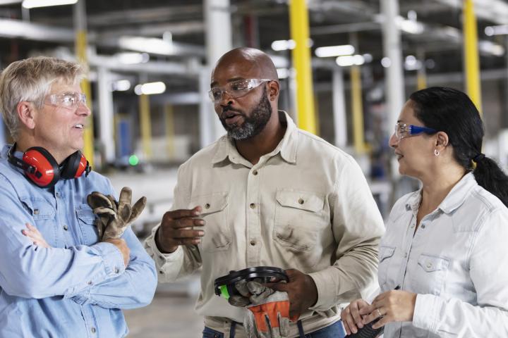 Two men and a woman having a conversation on a manufacturing floor, wearing safety glasses, ear protection and holding work gloves. 