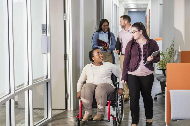 Colleagues chatting in the workplace; one is ambulating in a red wheelchair, others are walking.