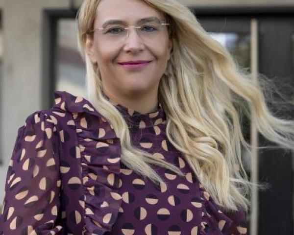 Maria Mexi is a woman with blonde hair and stands outside while wearing a maroon patterned top.