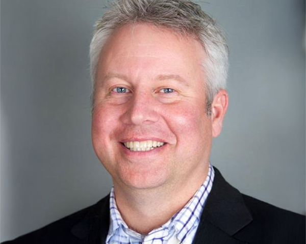 Smiling man with short gray hair wearing a black jacket over a light checkered shirt, posed against a neutral gray studio background.
