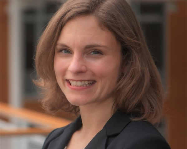 Smiling woman with shoulder-length light brown hair wearing a black blazer, standing indoors near a railing and windows.