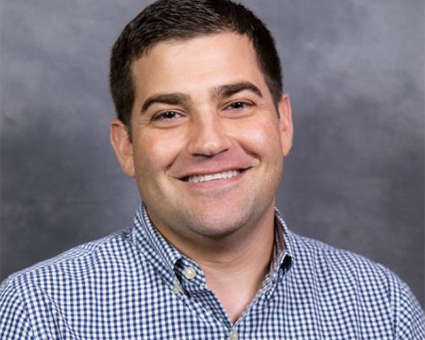 Smiling man with short dark hair wearing a blue-and-white checkered button-down shirt, posed against a neutral gray studio background.