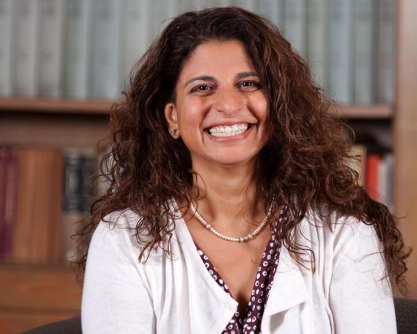 A smiling woman with curly hair sits in front of bookshelves, wearing a white cardigan and pearl necklace.