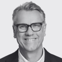 A black and white photo of a Colin Browne smiling as he wears glasses and has his short hair swooped over to his left, wearing a dark blazer and a light colored button-up shirt