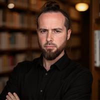 A man wearing a dark shirt stands arms crossed. He has a short cropped beard and dark hair. He stands in front of a bookshelf.