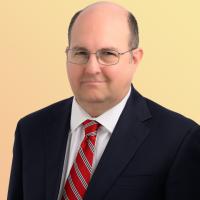 Stephen Vaugn stands wearing a dark suit with a white shirt and a red striped tie. He wears glasses and has hair on the side of his head.