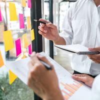 Two professionals review charts and brainstorm with colorful sticky notes on a glass wall during a collaborative planning session.