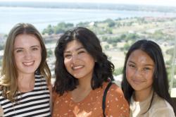 Three women smile into the camera with farm fields and a lake in the background