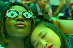 Two women sit close, one with eyes closed and her head resting on the shoulder of the second, whose glasses have paper eyes pasted to their lenses.